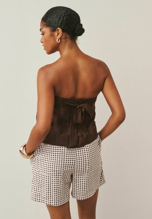 Woman with hair in a bun wearing gold hoop earrings, a strapless brown top with a back bow, and high-waisted brown-and-white checkered shorts.