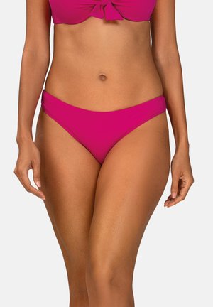 Kobieta w jasno-różowych majtkach od bikini i dopasowanym topie z przednim węzłem, pokazana od połowy tułowia do górnej części ud na jednolitym tle.