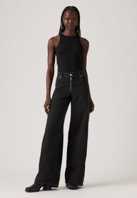 Jeans in denim nero a vita alta con una forma a gamba larga, dotati di chiusura con zip e bottone frontale, abbinati a un top aderente a coste nero.