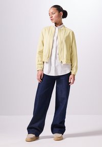 Giacca bomber gialla chiara con polsini elasticizzati, indossata sopra una camicia bianca a bottoni. Pantaloni jeans scuri a gamba larga e scarpe beige slip-on.