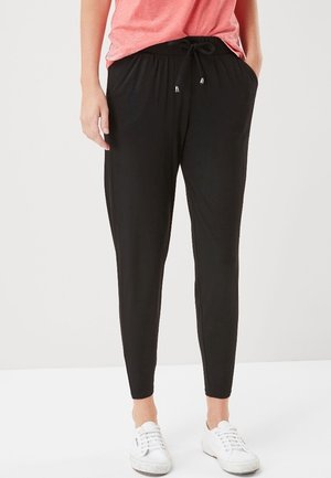 Persona que lleva pantalones negros con cordón, camiseta rosa de manga corta, con las manos en los bolsillos y zapatillas blancas, de pie sobre un fondo liso.