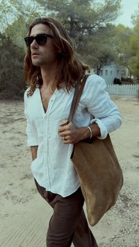 Vasarīga kamieļu krāsas suede plecu soma ar garu siksnu, kuru valkā cilvēks baltā pogājumā kreklā. Ārējā vide ar zaļām kokiem un taciņu.