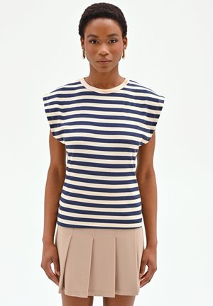 Femme aux cheveux courts portant des boucles d'oreilles créoles, un haut sans manches à rayures bleu marine et blanches, et une jupe plissée beige, debout devant un fond blanc.