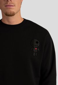 Primo piano di una persona che indossa una felpa nera con una piccola bambola nera ricamata con un cuore rosa sul lato sinistro del petto.