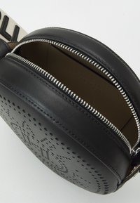 Rund svart läder crossbody-väska med silverblixtlås delvis öppen, perforerat logotypdesign fram och vit rem med svart text.