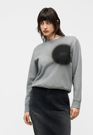 Młoda kobieta o ciemnych włosach nosi szarą bluzę z dużą czarną okrągłą naszywką na piersi oraz ciemną spódnicę.