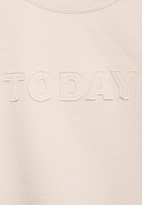 Svetlo bež pulover s posebno besedo "DANES" v teksturirani, vezenini pisavi po prsnici. Mehka, gladka tkanina s okroglim izrezom.