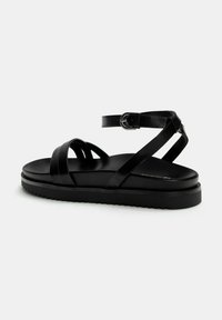 Chausson en cuir noir avec des lanières croisées, une sangle cheville réglable, une semelle plate et une semelle texturée pour une meilleure adhérence.