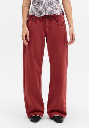 Personne portant un pantalon ample couleur rouille, des ballerines noires et une chemise à carreaux rentrée, debout devant un fond blanc.