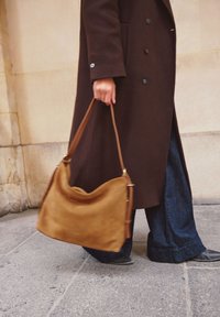 Brown suede shoulder bag with a smooth texture, rounded shape, and adjustable strap, carried alongside wide-leg denim pants on a stone surface.