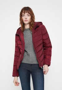 Bordo puffer jakna s kapuljačom, prošivenog dizajna, i srebrnim zatvaračem. Nosena preko sive pletene majice i uparena s tamnim trapericama.