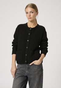 Schwarze, strukturierte Strickjacke mit rundem Hals, vorderen Knöpfen und zwei Taschen, kombiniert mit dunkelgrauen Jeans. Das Material wirkt dick und warm.
