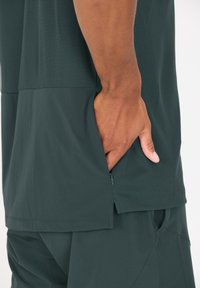 Donkergroen sportshirt met een gestructureerd bovenstuk en een smooth onderstuk, voorzien van een zijzak met rits en een relaxte pasvorm.