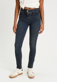 Hochtaillierte, dunkelblaue Skinny Jeans mit mittlerem Waschungsgrad, zwei Fronttaschen und Knopfverschluss, kombiniert mit weißen Sneakers.