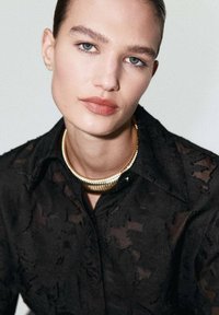 Camicia nera con texture e colletto, accompagnata da una collana d'oro spessa. Pelle liscia e trucco sottile con sopracciglia e labbra ben definite.