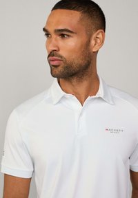 Weißes Polohemd mit kurzen Ärmeln und Kragen, mit roten Akzenten und dem "HACKETT SPORT" Logo in Grau auf der Brust. Glatte Textur.