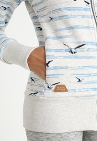 Sudadera con cremallera de color gris claro con un patrón de rayas azules y estampados de pájaros negros. Presenta puños acanalados y un parche de logo marrón en el lado.