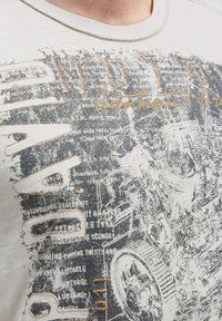 Helles graues T-Shirt mit einem strukturierten Grafikdruck und schwarzer Schrift sowie mechanischen Designelementen. Rundhalsausschnitt mit beigem Nähten.