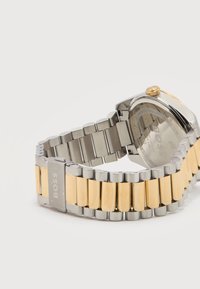 Ασημένιο και χρυσό μεταλλικό ρολόι χειρός με λογότυπο BOSS στην αγκράφα, εμφανίζοντας την πίσω όψη της θήκης του ρολογιού και το ανοιχτό μπρασελέ.