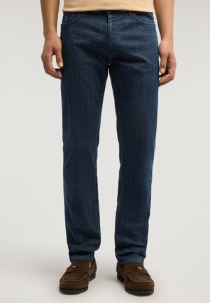 Uomo che indossa jeans blu scuro a taglio dritto e mocassini in camoscio marroni, in piedi contro uno sfondo chiaro e semplice.