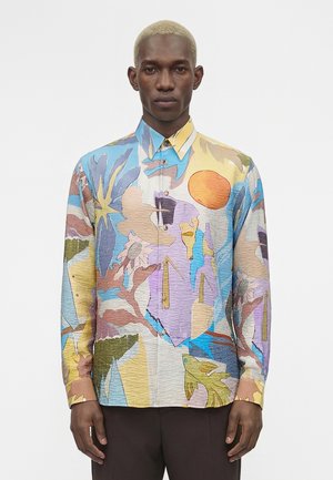 Muž s krátkymi blond vlasmi oblečený v viacfarebnej abstraktnej vzorovanej košeli na gombíky a tmavých nohaviciach stojaci pred jednofarebným pozadím.