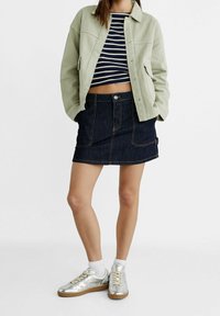 Kobieta w jasnozielonej kurtce, granatowej i białej pasiasty bluzce na krótki rękaw, ciemnoniebieskiej spódnicy dżinsowej, białych skarpetkach i srebrnych trampkach na białym tle.