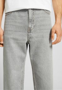 Jeans de mezclilla gris claro con un corte recto, costura visible y un acabado texturizado. Presenta una cinturilla estándar con bolsillos tradicionales.
