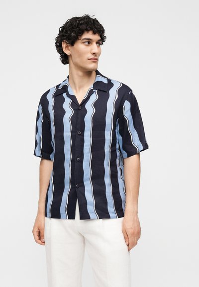 Άνδρας που φορά μαύρο, μπλε και λευκό κάθετο ριγέ πουκάμισο με κοντά μανίκια και λευκό παντελόνι, κοιτάζοντας προς τα αριστερά με φόντο μονόχρωμο.