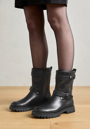 Fekete bokacsizmák sima bőr felülettel és texturált hálós felsőrésszel. Két hevederrel rendelkezik, kör alakú csattal. Barázdált gumitalp.