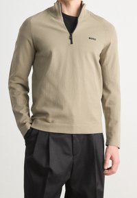 Man med beige kvartdraget tröja med "BOSS"-logo på vänstra bröstet och svarta plisserade byxor, stående med en hand i fickan.