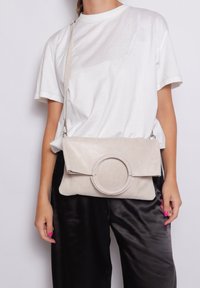 Borsa a tracolla beige con texture, con un accento circolare, esterno liscio e tracolla regolabile, tenuta contro uno sfondo neutro e indossata con una maglietta bianca.