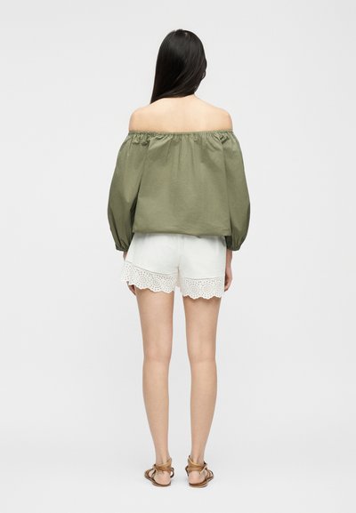 Γυναίκα που στέκεται με την πλάτη στην κάμερα, φορώντας πράσινη μπλούζα με έναν ώμο εκτός και λευκά σορτς με δαντέλα eyelet, καθώς και καφέ σανδάλια με δέσιμο στον αστράγαλο, σε λευκό φόντο.