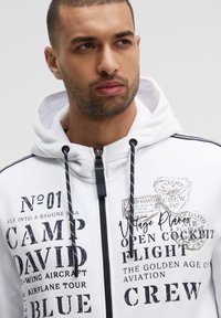 Sudadera blanca con cremallera y detalles en azul marino, que presenta gráficos impresos, incluyendo "CAMP DAVID" y visuales de aviones vintage. Cuerdas negras.