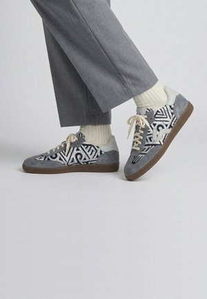 Personne portant un pantalon gris, des chaussettes crème et des baskets grises avec des motifs abstraits noirs et blancs et des semelles marron.