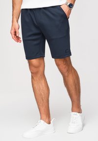 Homme portant un short bleu marine jusqu'aux genoux et des baskets blanches, debout avec une main dans la poche devant un fond uni.