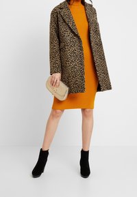 Płaszcz w panterkę nałożony na pomarańczową sukienkę z prążkami, w połączeniu z beżową torebką kopertową i czarnymi botkami na obcasie.