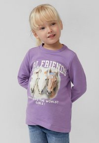 Meisje met blond haar dat een paarse sweatshirt draagt met daarop drie paarden en de tekst "Echte Vrienden" en "Wie bestuurt de wereld? Meisjes!"