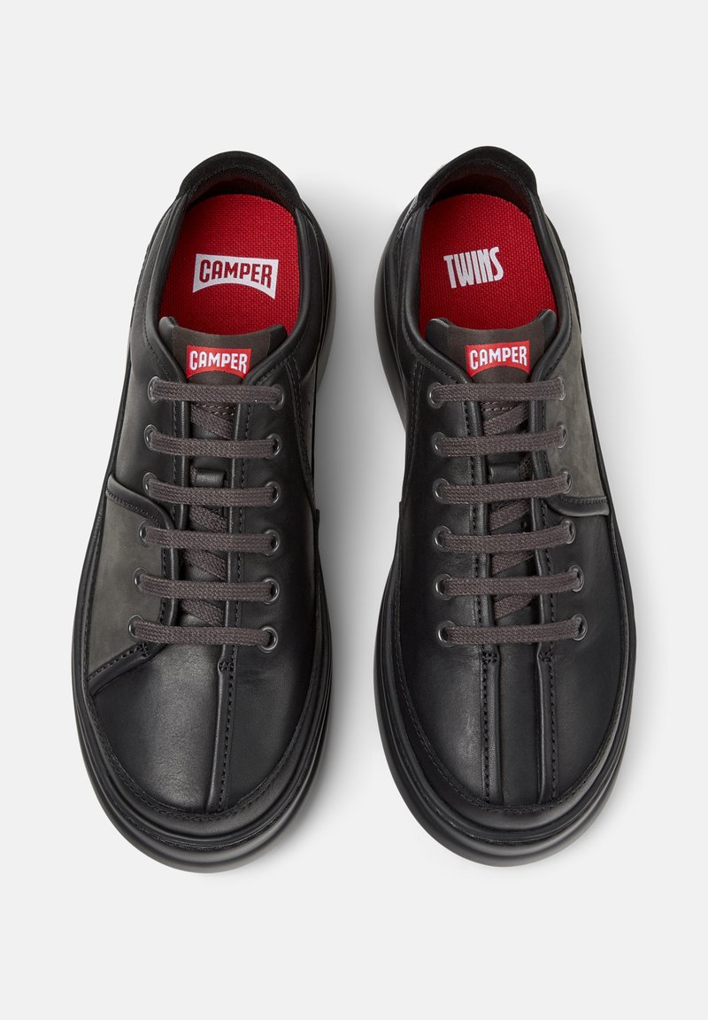 Schwarze Sneakers mit grauen Schnürsenkeln und einer roten Innensohle, die die Aufschriften "CAMPer" und "TWINS" trägt, von oben auf einem weißen Hintergrund betrachtet.