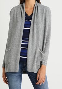 Kvinna som bär en grå öppen cardigan med fickor över en blårandig topp och blå jeans, stående med ena handen avslappnad.