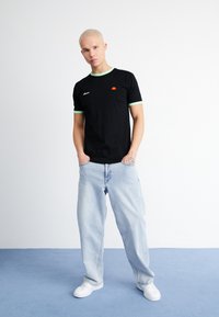 Black t-shirt with green collar accents and small logo on the chest, paired with light blue baggy jeans and white sneakers.