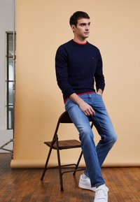 Pull en laine marine à col rond superposé sur une chemise rouge, associé à un pantalon en jeans bleus et des baskets blanches. Modèle assis sur une chaise en bois devant un fond uni.