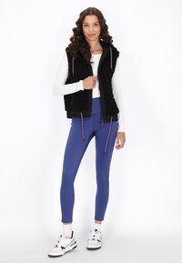 Schwarwolle, flauschige Weste mit Reißverschluss vorne und Kapuze, über einem weißen Oberteil und blauen Leggings. Getragen mit weißen Sneakers mit schwarzen Akzenten.