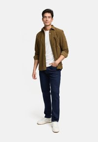 Camisa de botonada de pana marrón sobre una camiseta blanca, combinada con jeans azul oscuro y zapatillas blancas. El fondo es blanco sencillo.