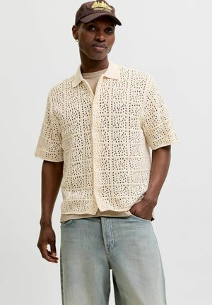 Mann trägt beige gehäkeltes kurzärmeliges Hemd mit Knöpfen, helle Jeans, beige T-Shirt darunter und braune Kappe mit goldener Stickerei.