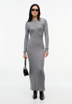 Femme portant une longue robe grise ajustée en maille torsadée, des bottines noires, des lunettes de soleil noires et tenant un sac à main noir.