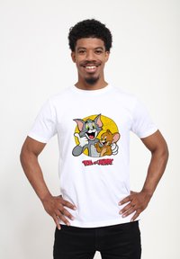 Bílá bavlněná tričko s barevným karikaturním motivem Toma a Jerryho na pozadí žlutého kruhu, s nápisem "TOM a JERRY" v tučném písmu pod ním.