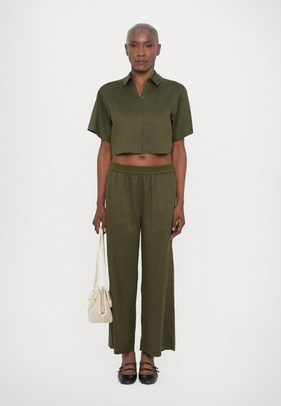Chemise cropped olive à manches courtes, associée à un pantalon à large plissé. Le mannequin tient un petit sac à main couleur crème et porte des chaussures plates foncées.