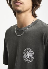 Grå bomulds t-shirt med korte ærmer og rund halsudskæring. Har et cirkulært hvidt grafisk print på brystet og en sølvfarvet kædeørering.