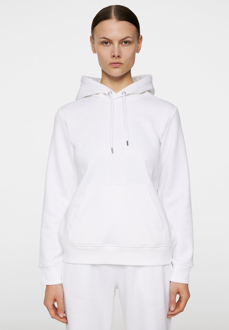Vit hoodie med en framficka, dragsnöre vid huvan och ribbade ärmslut. Tillverkad av mjukt tyg med minimal branding. Slät textur, avslappnad design.