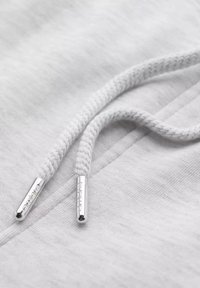 Tejido de sudadera blanca con superficie texturizada. Dos cordones blancos con extremos metálicos plateados, que presentan el nombre de la marca grabado.